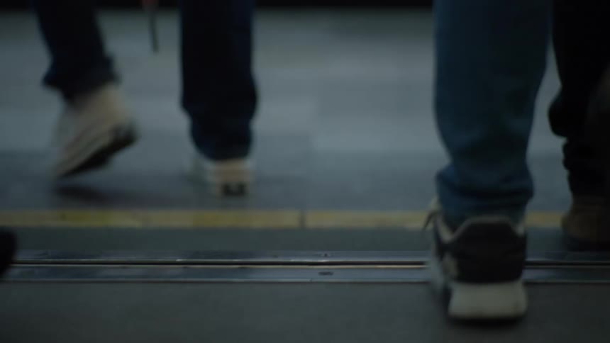 A slow-motion, low-angle view shows the feet of a crowd of commuters stepping off a subway train onto the platform as the doors open