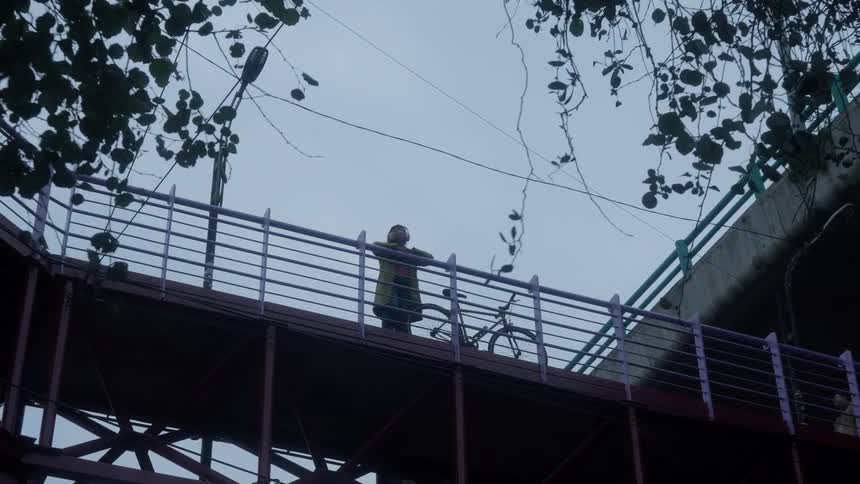 A thoughtful person with a bicycle stands on an urban pedestrian bridge on a gloomy, overcast day as the camera slowly zooms out