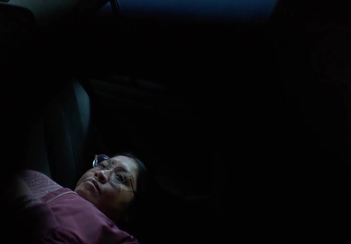 An exhausted woman with glasses lies down in the back of a car at night, with streetlights flashing across her face as she tries to sleep
