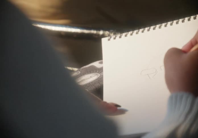 Over-the-shoulder view of a creative person with headphones sketching with a pencil in a notebook while commuting on a bus