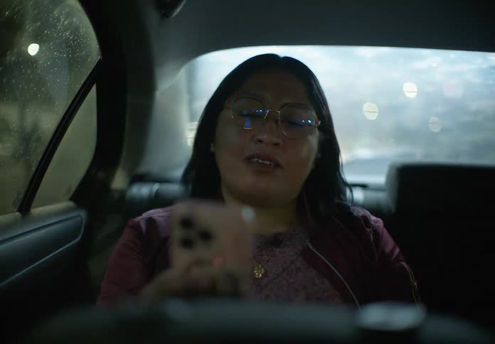 A woman wearing glasses smiles while looking at her smartphone in the backseat of a moving car during a rainy night