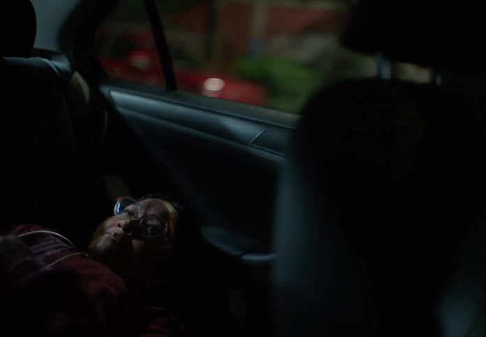 A tired woman with glasses lies down in the back of a moving car and falls asleep during a nighttime city ride