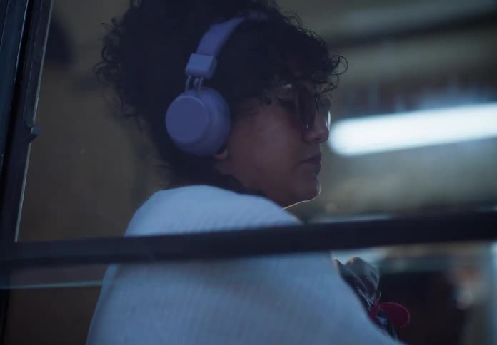A cinematic slow-motion view through a bus window of a curly-haired woman with headphones listening to music during her daily commute