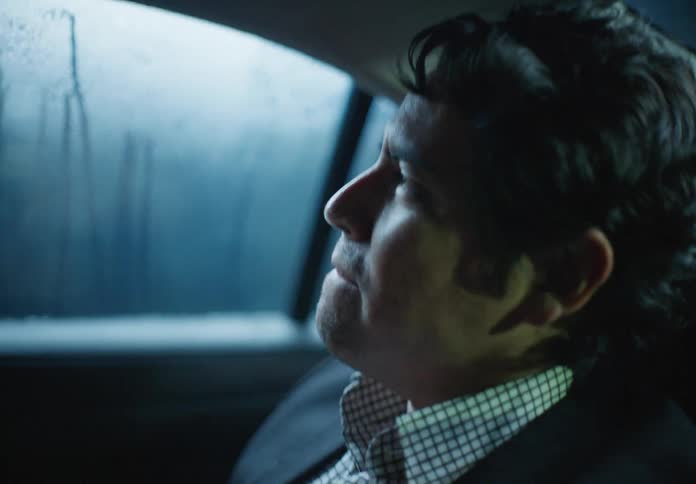 A pensive man in a business suit rides in the backseat of a car at night, watching the city lights pass by the rainy window
