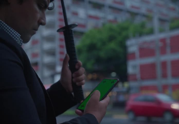 A pensive businessman holds an umbrella while checking his green screen smartphone on a rainy city street