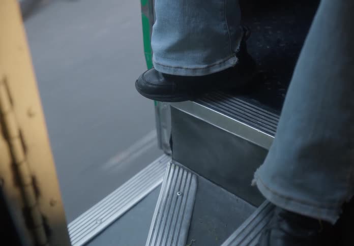 A high-angle view of a person's feet in jeans and black shoes as they ride on the steps of a moving bus, with the pavement speeding by outside