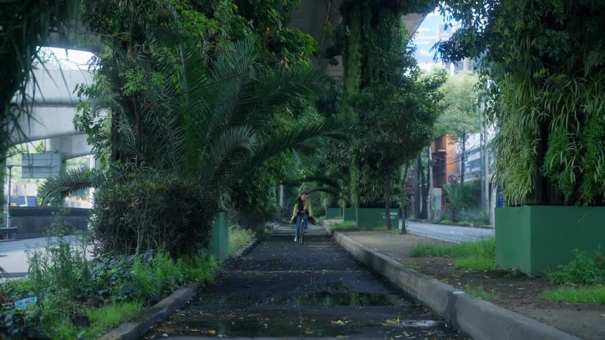 Slow motion shot of a woman wearing headphones as she rides her bicycle down a lush green path underneath a city highway