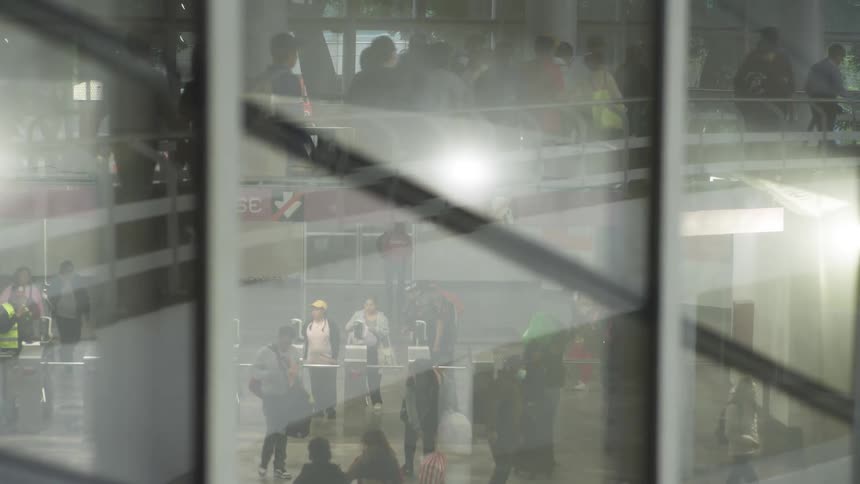 Reflections on a glass window show a bustling crowd of anonymous commuters moving through a sunlit public transit station