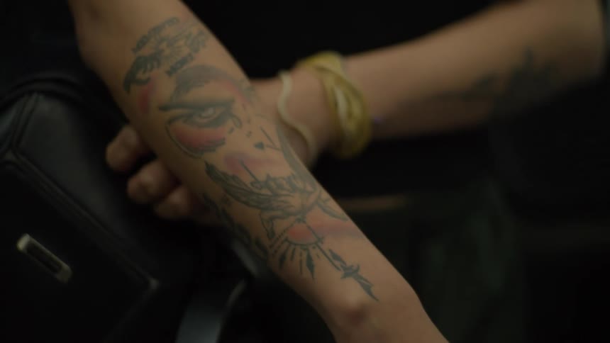 A close-up shot focuses on a person's forearm, showcasing intricate tattoos of an eye and the phrase 'Memento Mori'
