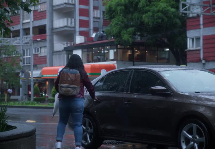 A woman with a backpack and clear umbrella walks through the rain-soaked city streets before hailing and getting into a waiting car