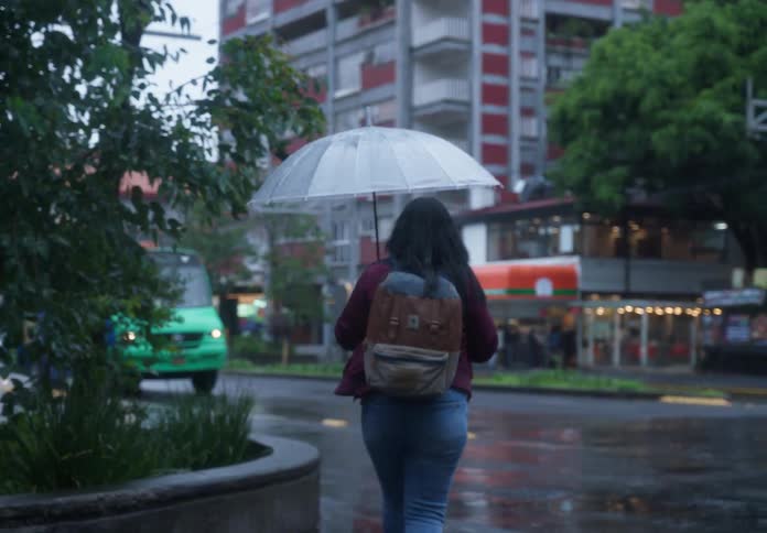 A woman with glasses and a backpack walks under a clear umbrella on a rainy city street, stopping to wait for traffic at a busy intersection