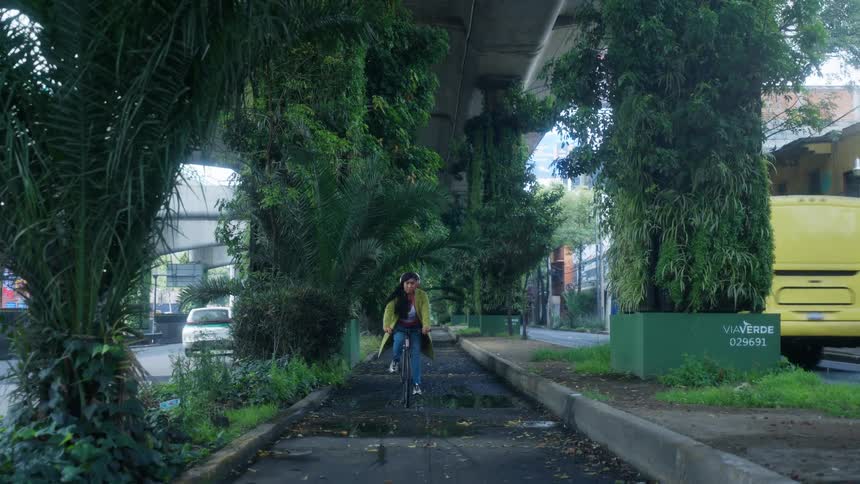 A young woman with headphones rides her bicycle along a unique urban greenway built beneath a highway overpass with plant-covered pillars