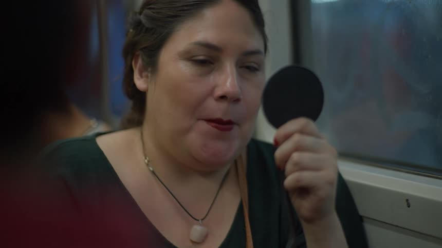 A woman carefully applies red lipstick and eyeshadow with a handheld mirror while commuting on the train in slow motion