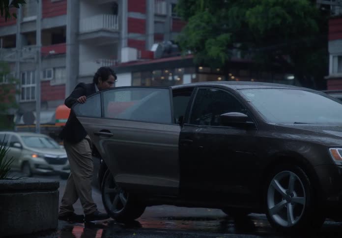 A man with an umbrella hails and gets into a car on a rainy city street with traffic passing by
