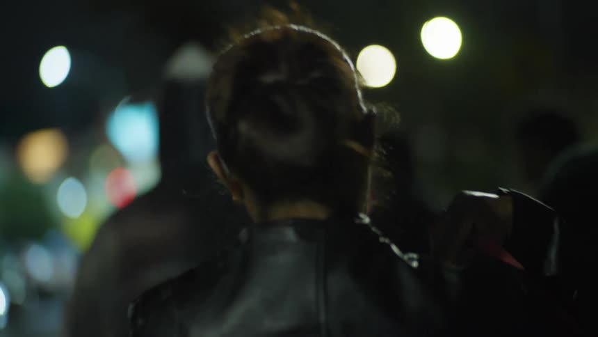 A person puts on their jacket and walks through the city at night, seen from behind in slow motion with cinematic bokeh lights