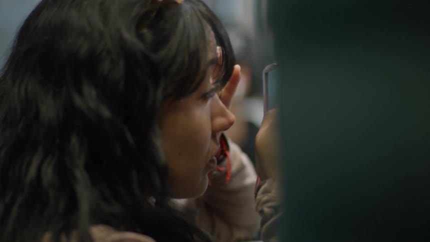 A close-up side profile of a young woman with dark hair applying makeup while looking in a small, pink, foldable mirror