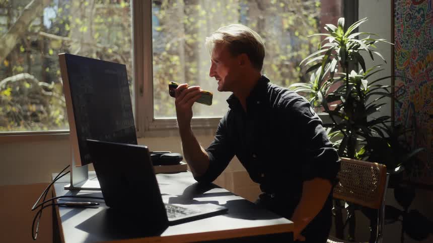 A smiling man enjoys an avocado toast while working on his dual-monitor setup in a bright, sunlit home office