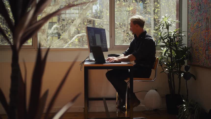 A man works on his desktop computer and laptop in a stylish, sunlit home office filled with plants