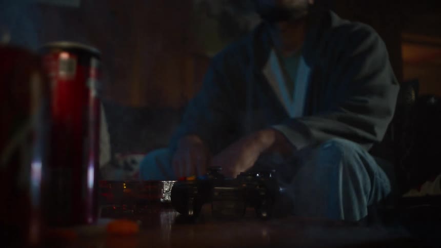 A frustrated man playing video games angrily slams his controller down on a table in a dark, smoky room