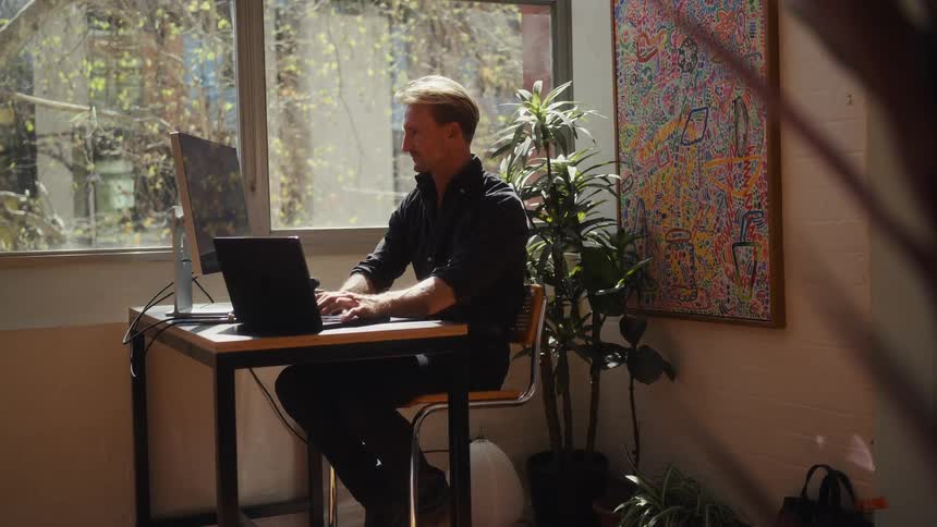 A focused man types on his laptop at a desk in a sunny, artistic home office