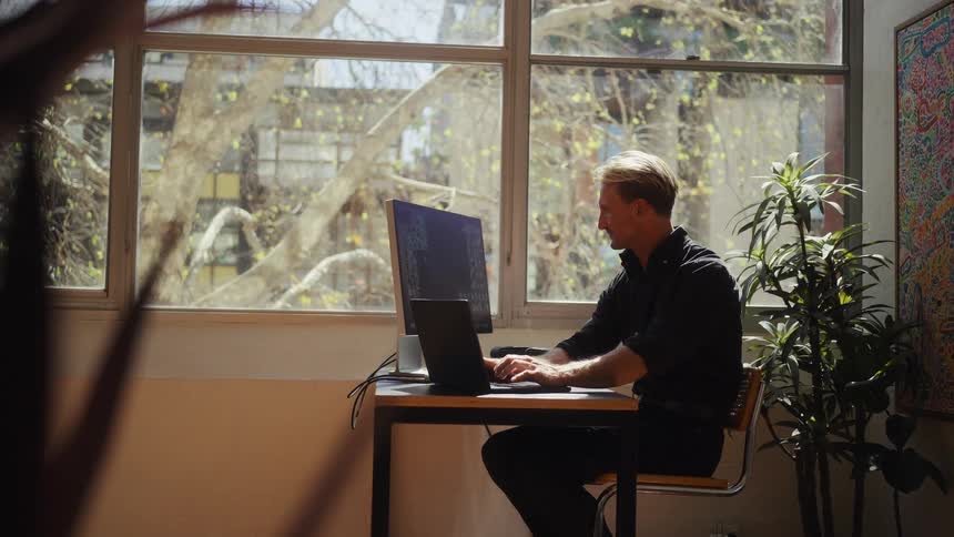 A focused man works diligently on his computer and laptop at his desk in a sunny, plant-filled home office