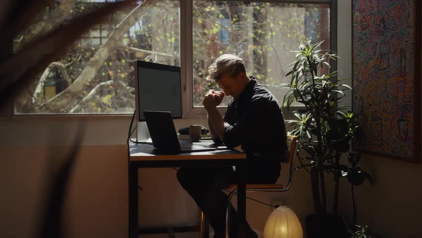 A thoughtful man working on his laptop in a sunlit home office with a slow zoom-in