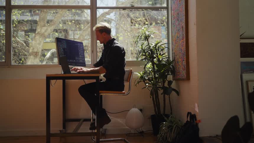 A focused man works diligently on his laptop at a high desk in a sunny, modern loft-style room