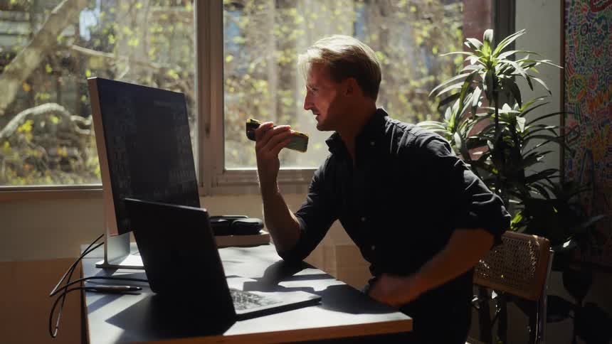 A professional man enjoys a piece of toast while working on his dual-screen computer setup in a brightly sunlit home office