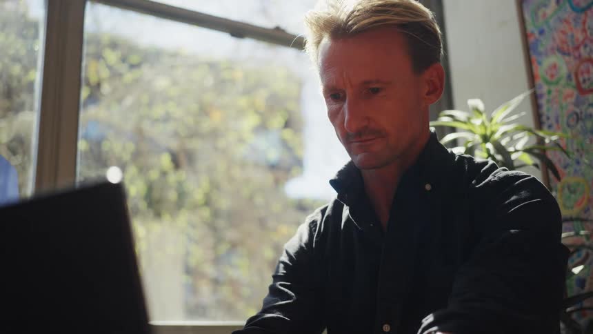 A focused man works on his laptop in a sunlit room before pausing to look thoughtfully out the window