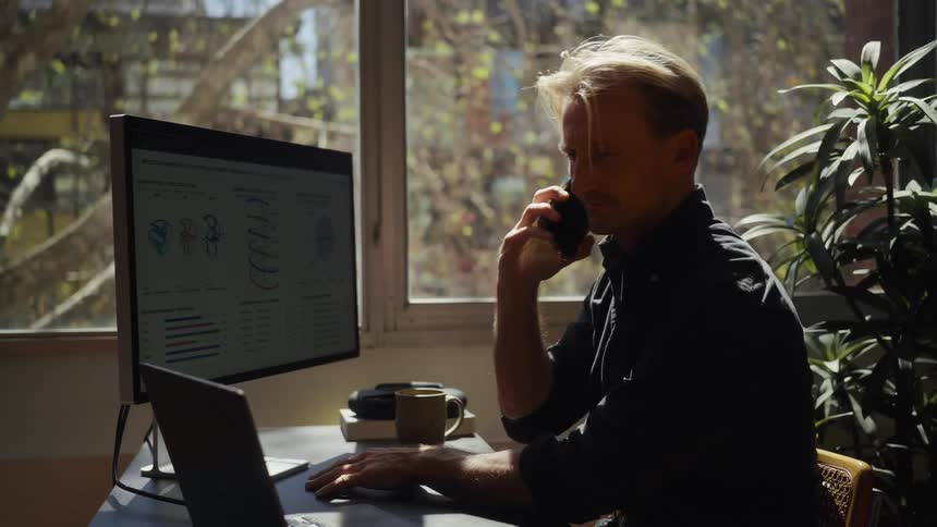 A focused businessman takes a call and works on his laptop and monitor, analyzing charts and data in a bright, modern home office