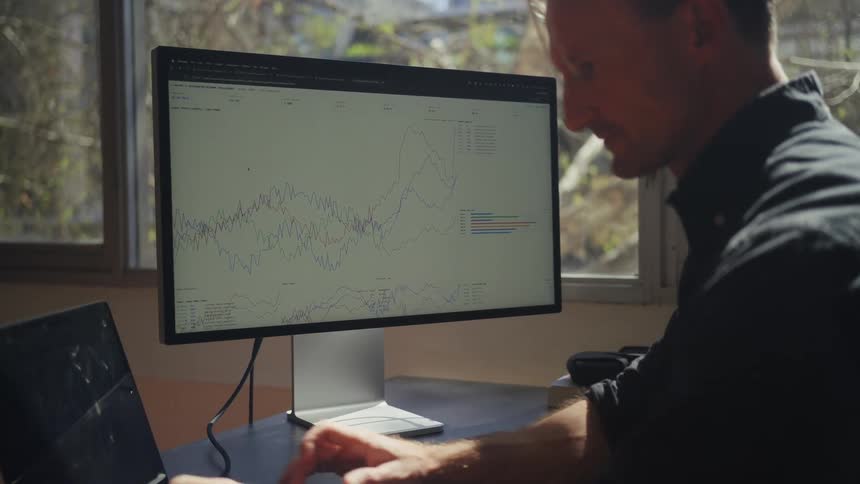 A man sits at his desk in a sunny office, closely analyzing complex data charts and graphs on his large computer monitor