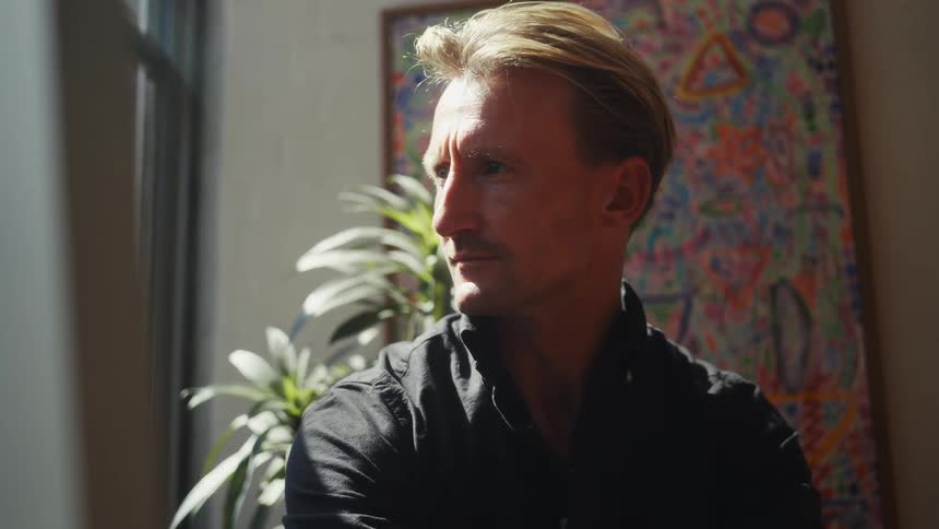 Sunlight illuminates the face of a pensive man as he gazes thoughtfully out a window in a quiet room