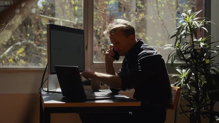 A businessman multitasks on a phone call while pointing at his laptop screen in a sunlit home office with a large window