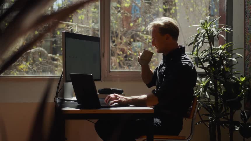 Slow motion side view of a focused man working on his laptop and drinking coffee at his desk by a sunny window