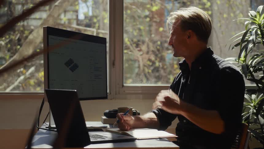 A focused man works from his sunlit home office, analyzing data on his computer and taking notes in his notebook