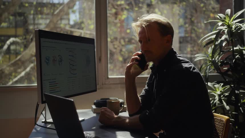 A professional man talks on the phone while analyzing complex data and charts on his laptop and large monitor in a sunlit office