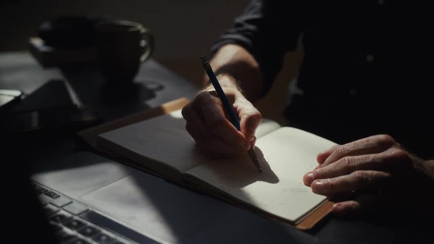 A close-up shot captures a person's hands diligently writing and sketching in a notebook, illuminated by a dramatic shaft of sunlight