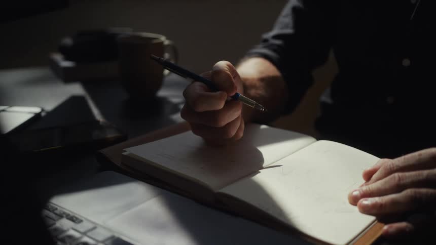 A man sketches ideas in a sun-drenched notebook at his desk in a dimly lit room