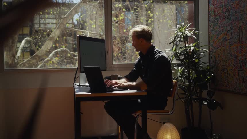 A professional man works diligently on his laptop at a desk bathed in natural light from a large window in his home office