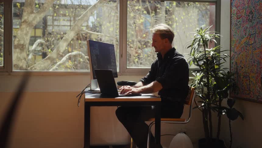 A professional works diligently on his laptop and vertical monitor in a sun-filled, artistic home office