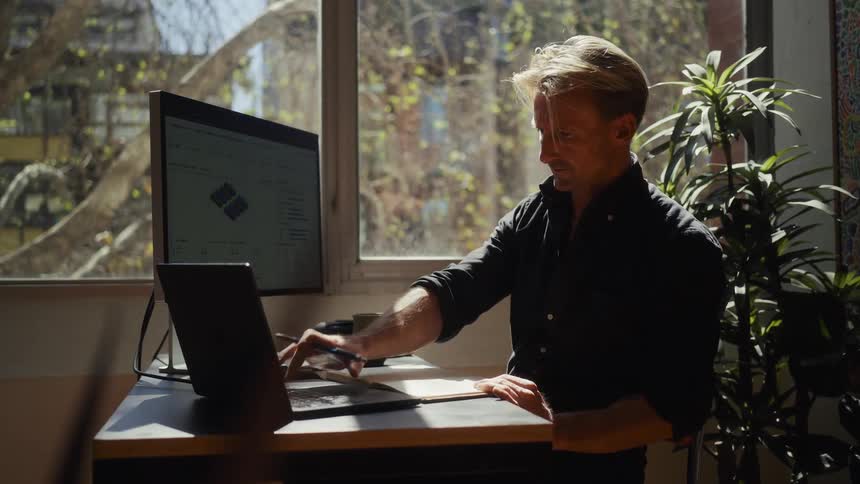 A focused man works at his standing desk in a sunlit home office, writing notes and using his laptop and computer monitor