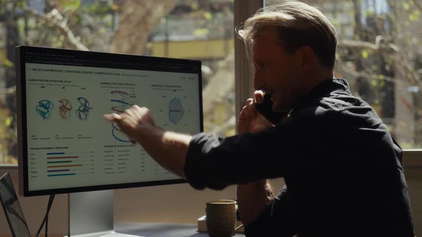 A male scientist on a phone call discusses complex data on his computer, pointing at models of human protein and genomic structures