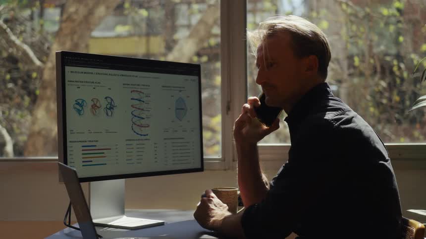 A male scientist or researcher analyzes complex molecular data on his computer while on a phone call in his sunlit office