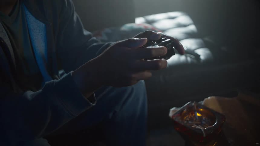 A gamer's hands grip a controller while playing a video game in a dark, smoky room with an ashtray full of cigarettes on the table