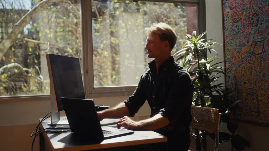 A focused man sends a voice message and works on his laptop and monitor in a sunlit home office