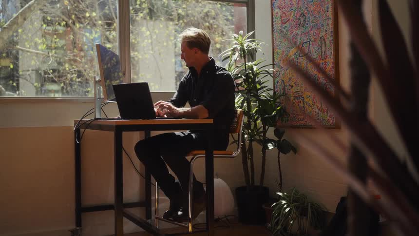 A man works on a laptop at a high desk in his sun-drenched home office with a slow push-in camera movement