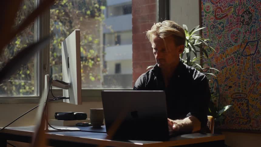 Slow dolly reveal of a focused man working on his laptop and desktop computer in a sunlit home office