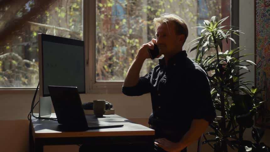 A professional man takes a pleasant phone call before continuing his work on a laptop in a bright, sunlit home office