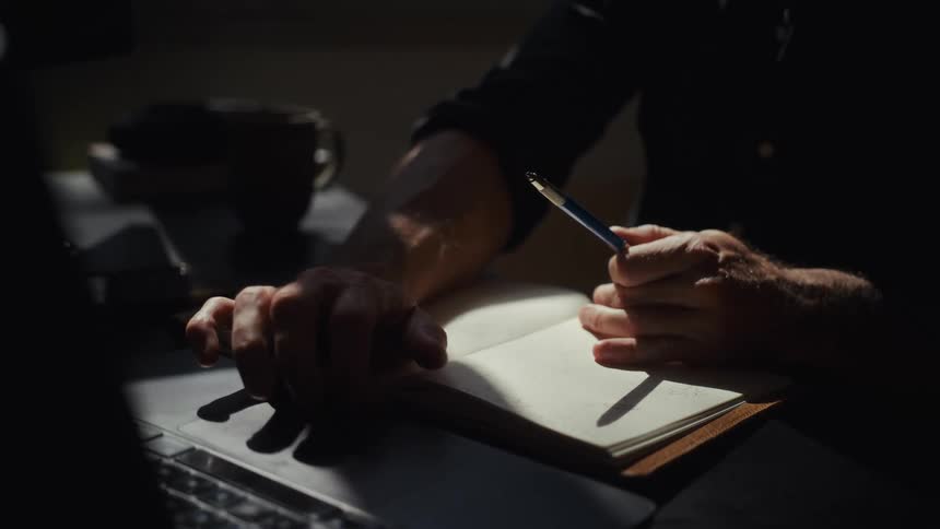 A thoughtful man sits at a dimly lit desk, using his laptop and holding a pen over a notebook