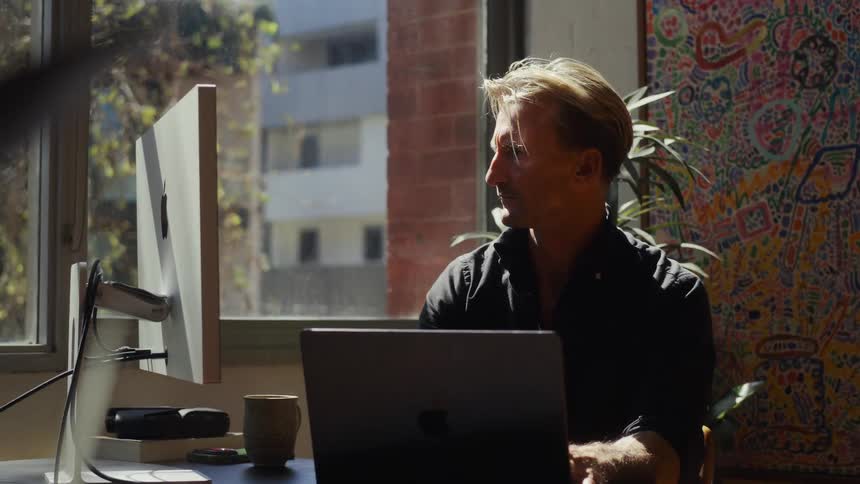 A man works from home on his laptop at a desk in a creative, sunlit office space
